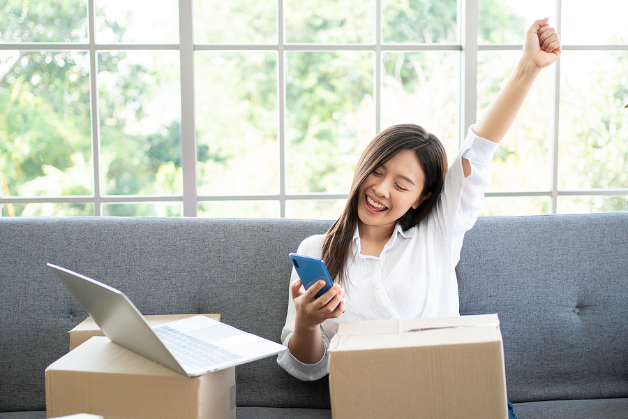 Happy Asian woman with moving boxes and laptop in sunny living room celebrating she has found the best flat fee MLS company on her cellphone.