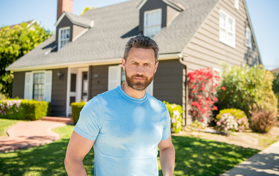 Male homeowner in tee shirt in front of his neat two story family home deciding if he will rent or sell the home